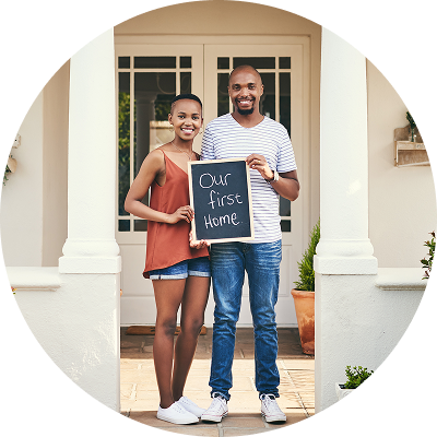 First-time homebuyer couple standing on their new porch, smiling and holding a sign that reads 'Our First Home.