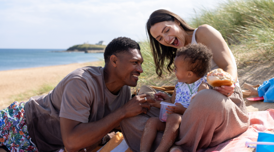 The young parents sit on a colourful blanket with their toddler, surrounded by beach toys and picnic items.