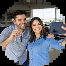 A smiling couple taking a selfie in front of their new car at a dealership, celebrating their first car purchase.