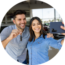 A smiling couple taking a selfie in front of their new car at a dealership, celebrating their first car purchase.