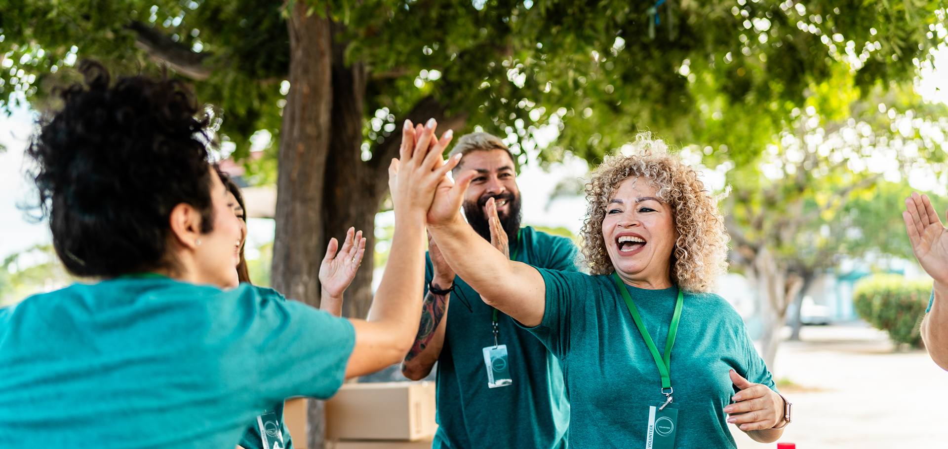 A diverse group of volunteers wearing matching green shirts smile and high-five each other outdoors under leafy trees.