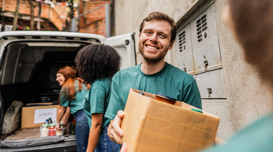 A smiling volunteer in a green shirt carries a cardboard box while other volunteers unload supplies from a van in an urban alley.