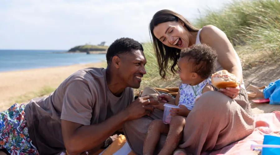 The young parents sit on a colourful blanket with their toddler, surrounded by beach toys and picnic items.