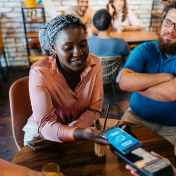 Young group of friends relaxing in a café, drinking juice and coffee. Young woman paying the bill via contactless channel by mobile banking application.