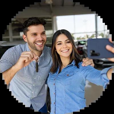 A smiling couple taking a selfie in front of their new car at a dealership, celebrating their first car purchase.