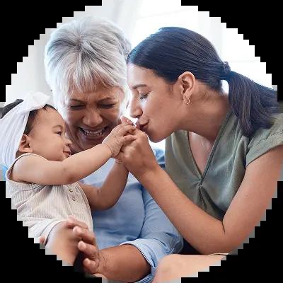 A mother kisses her baby’s hand while sitting on her grandmother’s lap, symbolizing generations—reminder to update your account beneficiaries.