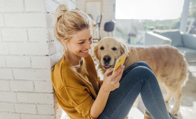 A woman sits on the floor with her dog by her side, checking the growth of her Civic Choice Certificate account on her phone.
